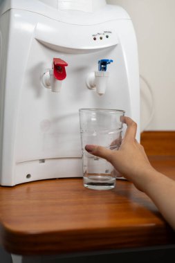 Bir el, temiz bir bardağa, ahşap bir yüzeye yerleştirilmiş su dağıtıcısı kullanarak temiz su döker..