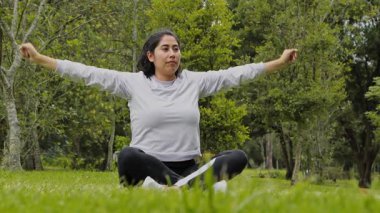 Bir kadın parkta vücudunu esnetirken derin nefes alıyor. Rahatlamayı, iyileşmeyi ve açık hava egzersizinden sonra dikkatli nefes almayı temsil ediyor..