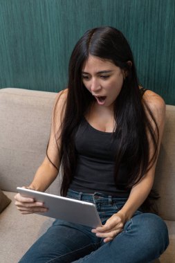 Kanepede oturan genç bir kadın ağzı açık bir tabletle ekrandaki beklenmedik içeriğe tepki veriyor, şok, duygu, dijital eğlence ve modern ev medya tüketimini temsil ediyor..