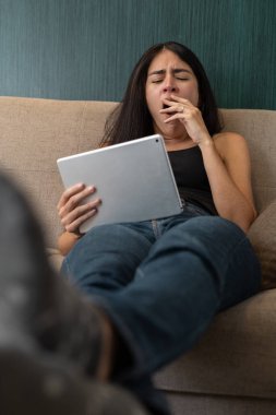Kanepede oturan genç bir kadın elinde tabletle uykusuzluktan esniyor, yorgunluk, yorgunluk ve akşamları evde dijital eğlence ile dinleniyor..
