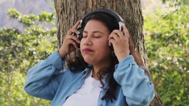 Genç bir kadın, kulaklıkları açık havada huzurlu müziğin keyfini çıkarıyor. Etrafı doğa ile çevrili, sakinliğini ve iç huzurunu ifade ediyor..