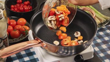 Bir kadın sıcak bir tavaya doğranmış domates, havuç ve mantar ekler. Parlak bir mutfak ortamında renkli ve sağlıklı bir ev yemeği pişirir..