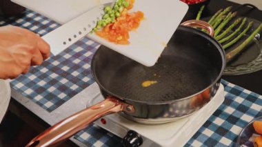 Doğranmış domatesleri ve kuşkonmazı bıçakla kesme tahtasından tavaya kaydırarak yemek pişirme, yemek hazırlama ve mutfak rutinini gösteren ellerinizi kapatın..