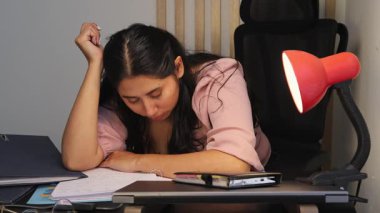 Yorgun genç bir kadın masasında kağıtları inceliyor, uyanık kalmaya çalışırken başını koluna yaslıyor, yorgunluğu, aşırı çalışmayı, stresi ve çalışma rutinini temsil ediyor..