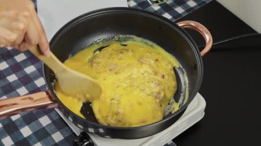 Bir kadın çırpılmış yumurta hazırlarken tavada yumurta taşıyor. Kahvaltı, ev yemeği ve günlük mutfak rutinini temsil ediyor..