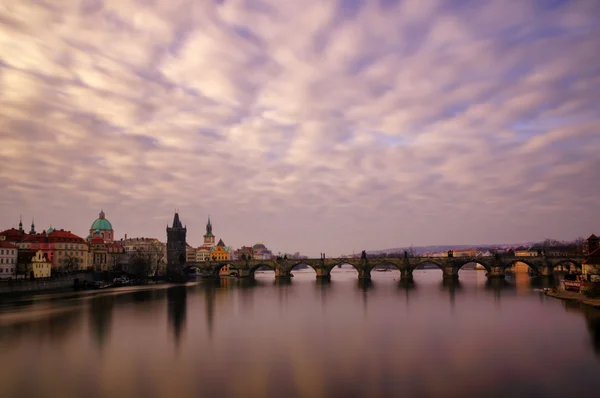 Belo nascer do sol perto da famosa Ponte Charles com céu nublado e reflexo de água. Ponte com torre incrível e estátuas antigas no início da manhã. Dia ensolarado típico em Praga, República Checa . — Fotografia de Stock