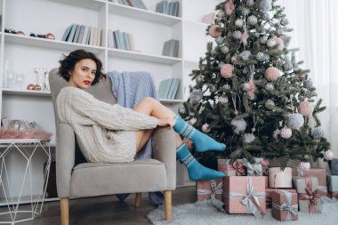 Güzel ve mutlu esmer, arkasında Noel ışıkları olan sıcak bir süveter giymiş sandalyede oturuyor.