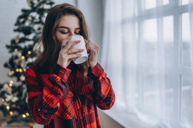Güzel esmer kız pencerenin yanında duruyor, kırmızı gömlek ve sıcak çoraplar giyiyor ve arkasında bir fincan kahve ve Noel ışıkları var.