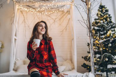 Yatakta güzel esmer kız kırmızı gömlekli ve sıcak çoraplı bir fincan kahve ve Noel ışıkları arkasında.