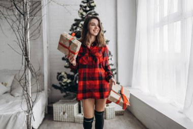 Noel, yeni yıl. Gömlekli ve çoraplı güzel bir kadın Noel hediyesi kutusuyla evde yürüyor, yılbaşı ağacı arkasında.