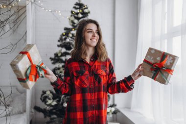 Noel, yeni yıl. Gömlekli ve çoraplı güzel bir kadın Noel hediyesi kutusuyla evde yürüyor, yılbaşı ağacı arkasında.