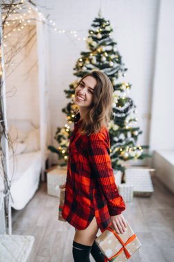 Noel, yeni yıl. Gömlekli ve çoraplı güzel bir kadın Noel hediyesi kutusuyla evde yürüyor, yılbaşı ağacı arkasında.