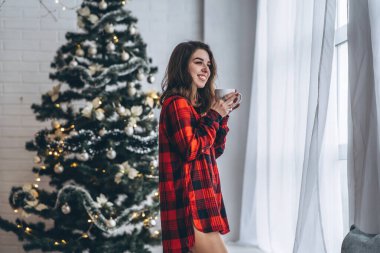Güzel esmer kız pencerenin yanında duruyor, kırmızı gömlek ve sıcak çoraplar giyiyor ve arkasında bir fincan kahve ve Noel ışıkları var.
