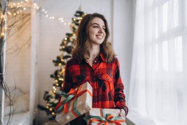 Noel, yeni yıl. Gömlekli ve çoraplı güzel bir kadın Noel hediyesi kutusuyla evde yürüyor, yılbaşı ağacı arkasında.