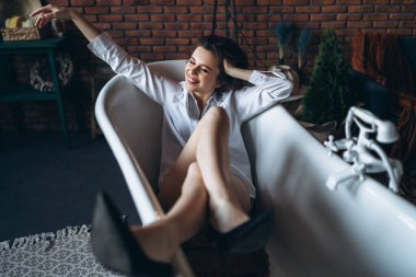 Güzel bir esmerin portresi, boş bir banyoda uzanmış, bacakları havada, beyaz gömlek ve siyah ayakkabılar giymiş, kırmızı tuğlalı arka planda..