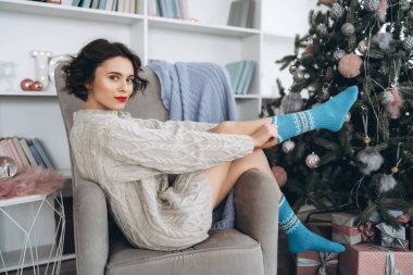 Güzel ve mutlu esmer, arkasında Noel ışıkları olan sıcak bir süveter giymiş sandalyede oturuyor.