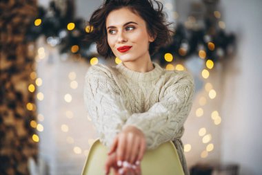 Güzel ve mutlu esmer, arkasında Noel ışıkları olan sıcak bir süveter giymiş sandalyede oturuyor.