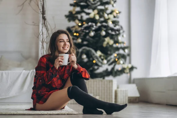 かなりブルネットの女の子座っている床に赤いシャツと暖かい靴下でコーヒーとクリスマスツリーの後ろ — ストック写真