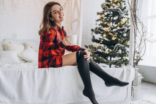 かわいいですブルネット女の子でベッド身に着けています赤いシャツと暖かい靴下とともにコーヒーとクリスマスライトBehin — ストック写真