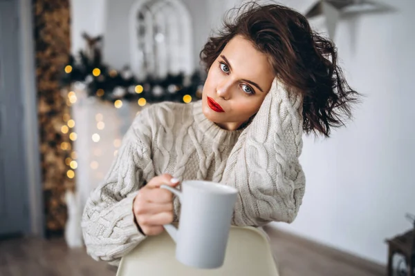 漂亮而快乐的黑发姑娘 穿着暖和的毛衣坐在椅子上 手里拿着一杯带圣诞灯的热饮料 — 图库照片