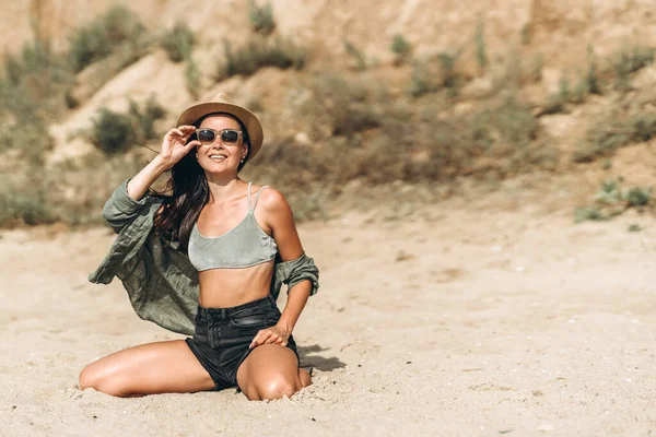 Şapkalı esmer kız plajda dinleniyor ve arka planda kayalar var.