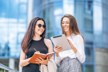 Tabletli ve defterli iki genç iş kadını modern iş merkezinin yakınlarında yürüyorlar.