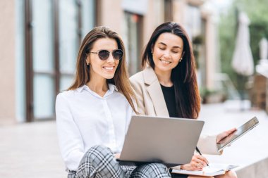 Tablet ve dizüstü bilgisayarlı iki genç iş kadını modern iş merkezinin yanında oturuyor.