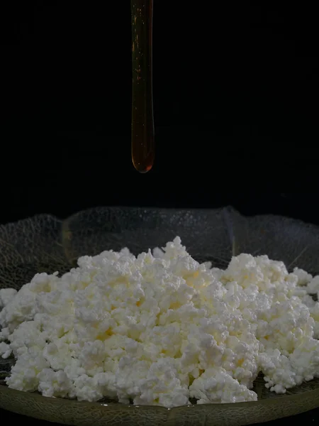 白いチーズをガラス皿に砕き 黒の背景に蜂蜜を注ぐ — ストック写真