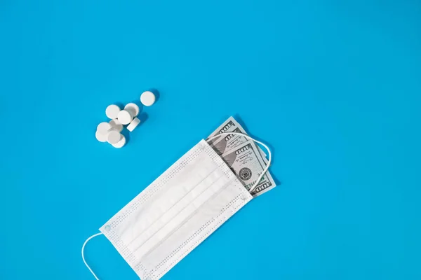 Pills and medical face mask covered one hundred dollar bill on blue background. Concept of economic crisis caused by coronavirus pandemic, expensive treatment and vaccine, the pharmaceutical business.