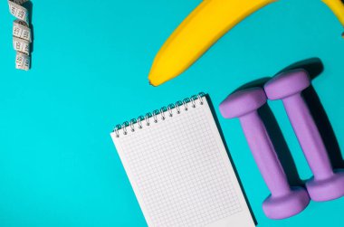 Mor halterler, muz, not defteri ve mavi arka planda mezura. Üst Manzara. Fitness konsepti, kilo kaybı, diyet.