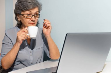 Yaşlı kadın kahve içiyor ve dizüstü bilgisayara şaşırmış görünüyor..