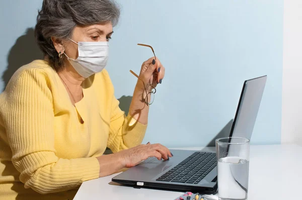 Evde dizüstü bilgisayar kullanan gözlüklü ve tıbbi maskeli yaşlı bir kadın. Doktorla çevrimiçi danışma, internet araması, kişisel izolasyon, yeni normal.