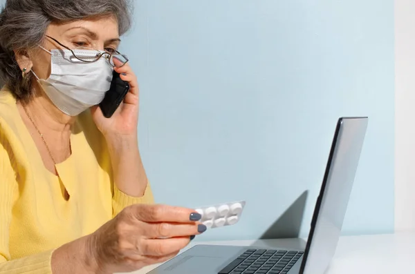 Senior woman with glasses and medical mask reading instructions for the medicine, copy space. Elderly woman using laptop at home. Concept for online consultation with doctor, new normal.