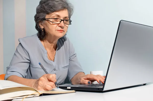 Laptop kullanan ve günlük yazan yaşlı bir kadın. Seyahat, serbest çalışma, emeklilik kavramı.