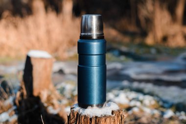 Termos kütüğün üzerinde duruyor, yakın plan. Kış ormanlarında yürüyüş için kullanılan vakum mavisi matara. Seyahat konsepti, macera, sıcak kış içeceği.