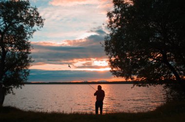 Gölde gün batımında balık tutmak. Balıkçı bir olta atar, arkadan bakar. Gölün üzerinde gün batımı manzarası ve uçan bir martı..