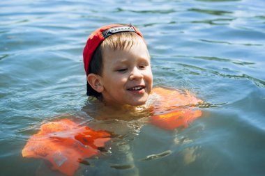 Doğada, açık havada çocuk eğlencesi. Şapkalı ve şişme kolluklu mutlu küçük çocuk suda yüzüyor. Çocuk yüzmeyi öğrendi..