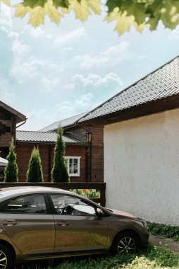 Bir kır evi manzarası, bahçedeki park edilmiş bir araba bir ağacın altında ahşap bir evin arka planında. Mavi gökyüzü ve yeşil yapraklar. Banliyö yaşamında huzur dolu bir huzur.