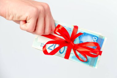 Bir adamın eli, açık arkaplanda kırmızı bir kurdeleyle bağlanmış 200 yeni İsrail şekeli banknotlardan oluşan bir tomar tutuyor.