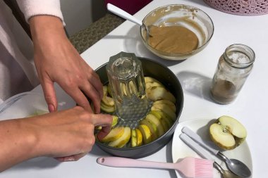 Bir kadın Charlotte 'un pişirme kabına dilimlenmiş elma koyar. Yakınlarda elmalı turta yapmak için diğer malzemeler ve araçlar var..