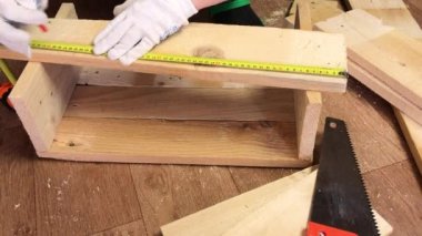 Adam boyutları mezurayla ölçüyor ve tahtalardaki kesme çizgilerini işaretliyor. Sebze toplamak için bir depolama kutusu yapıyor. Bir apartman dairesinde salgın sırasında sebze toplamak.