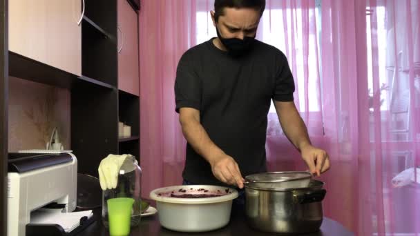 Чоловік у медичній масці фільтрує ферментовану м'якоть винограду через сито. Виготовлення домашнього вина під час епідемії . — стокове відео