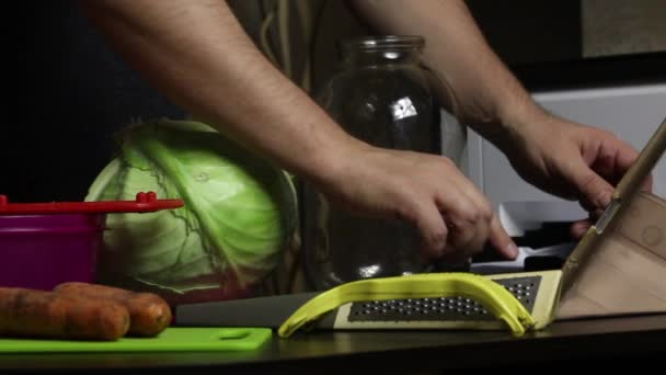 एक माणूस व्हिडिओ लिंकद्वारे केबज चिरण्याची प्रक्रिया प्रसारित करतो. घरात सॉरक्रॉट. त्याच्या शेजारील टेबलावर कोबी, गाजर आणि स्वयंपाकाची साधने आहेत. बंद-अप शॉट . — स्टॉक व्हिडिओ