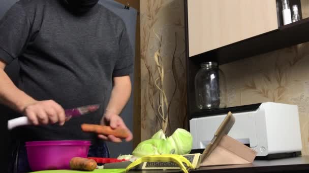 Um homem com uma máscara médica transmite o processo de descascar cenouras via link de vídeo. Chucrute em casa durante uma pandemia. — Vídeo de Stock