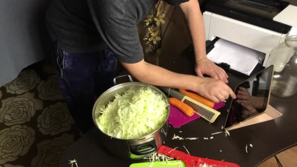 Ένας άντρας με ιατρική μάσκα τρίβει ένα καρότο. Δίπλα του είναι ψιλοκομμένο λάχανο σε μια κατσαρόλα. Μεταδίδει τη διαδικασία λείανσης μέσω σύνδεσης βίντεο. Sauerkraut στο σπίτι κατά τη διάρκεια μιας πανδημίας. — Αρχείο Βίντεο