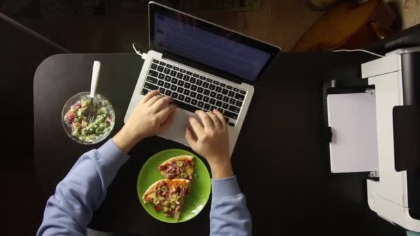 男がノートパソコンの後ろにテキストを入力している。テーブルの近くにはピザとサラダがあります。上から撮影. — ストック動画
