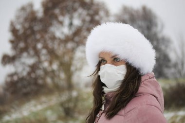 Parkta yürürken üzerinde kışlık giysiler olan bir kızın portresi. Yüzünde tıbbi maske var. Yüzün yarısı vurulmuş..