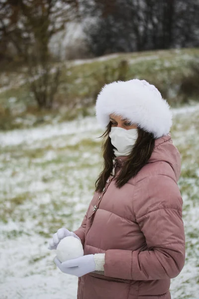 A girl in a medical mask for a walk. Holds a ball of snow in his palms. The ground is covered with the first snow