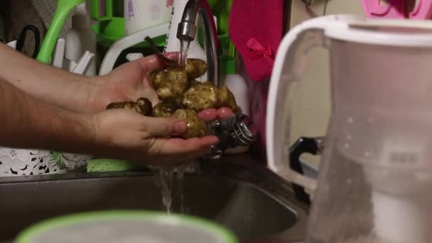 Un hombre lava la alcachofa sucia de Jerusalén. En el fregadero de la cocina bajo agua corriente. Cubre el agua y se limpia las manos con un trapo. — Vídeos de Stock