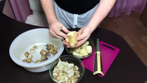 A man peels Jerusalem artichoke with a kitchen knife. Throws the peel into one container, the peeled vegetable into another. — Stock Video